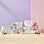 Le Creuset�s Sorbet Collection displayed with a pastel-striped backdrop. The image features colorful packaging for the collection, stacked bowls, mugs, and a footed ice cream bowl with a spoon. The products are arranged on pastel blocks in shades of pink, yellow, and lavender.