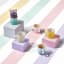 Le Creuset�s Sorbet Collection displayed with a pastel-striped backdrop. The image features colorful packaging for the collection, stacked bowls, mugs, and a footed ice cream bowl with a spoon. The products are arranged on pastel blocks in shades of pink, yellow, and lavender.