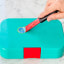 Dreamfarm Self Sharpening Cleaning Scrapers, Set of 2 removing a sticker on a lunchbox 