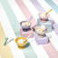 Le Creusetï¿½s Sorbet Collection displayed with a pastel-striped backdrop. The image features colorful packaging for the collection, stacked bowls, mugs, and a footed ice cream bowl with a spoon. The products are arranged on pastel blocks in shades of pink, yellow, and lavender.