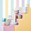Le Creusetï¿½s Sorbet Collection displayed with a pastel-striped backdrop. The image features colorful packaging for the collection, stacked bowls, mugs, and a footed ice cream bowl with a spoon. The products are arranged on pastel blocks in shades of pink, yellow, and lavender.