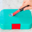 Dreamfarm Self Sharpening Cleaning Scrapers, Set of 2 removing a sticker on a lunchbox 