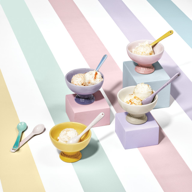 Le Creuset�s Sorbet Collection displayed with a pastel-striped backdrop. The image features colorful packaging for the collection, stacked bowls, mugs, and a footed ice cream bowl with a spoon. The products are arranged on pastel blocks in shades of pink, yellow, and lavender.