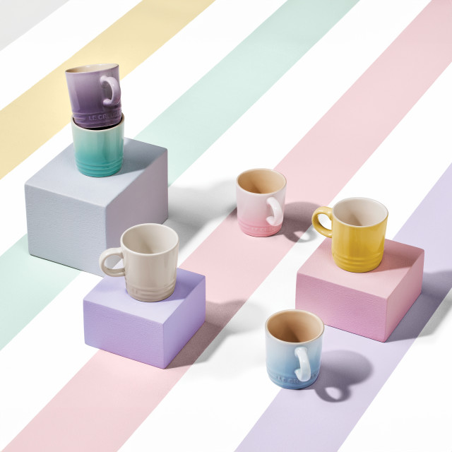 Le Creuset�s Sorbet Collection displayed with a pastel-striped backdrop. The image features colorful packaging for the collection, stacked bowls, mugs, and a footed ice cream bowl with a spoon. The products are arranged on pastel blocks in shades of pink, yellow, and lavender.