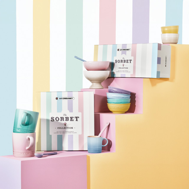 Le Creuset�s Sorbet Collection displayed with a pastel-striped backdrop. The image features colorful packaging for the collection, stacked bowls, mugs, and a footed ice cream bowl with a spoon. The products are arranged on pastel blocks in shades of pink, yellow, and lavender.