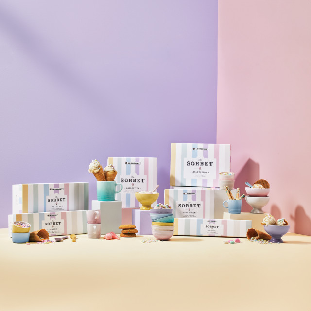 Le Creusetï¿½s Sorbet Collection displayed with a pastel-striped backdrop. The image features colorful packaging for the collection, stacked bowls, mugs, and a footed ice cream bowl with a spoon. The products are arranged on pastel blocks in shades of pink, yellow, and lavender.