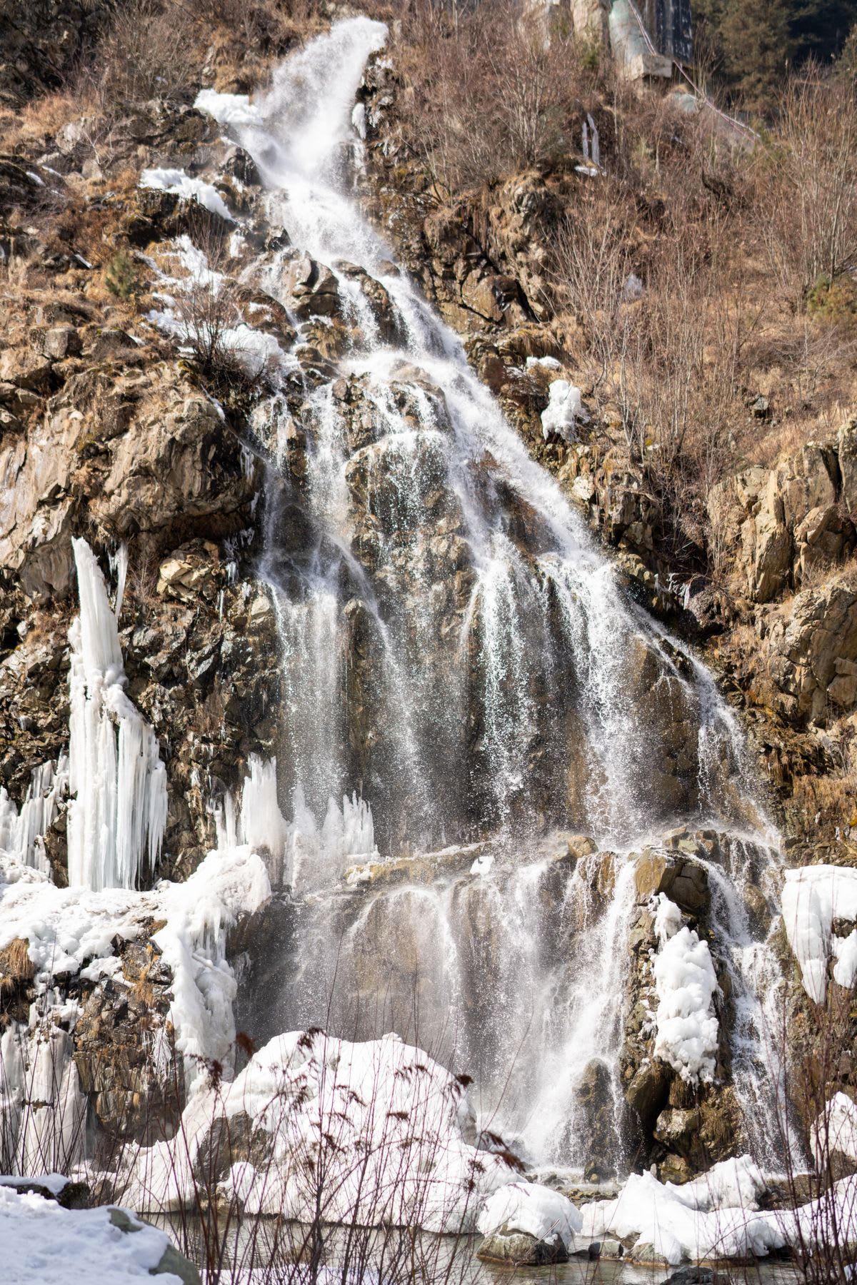 Drung waterfall above Tangmarg in September -- a single-thread cascade of approximately 20 metres dropping off a dark sandstone cliff face, surrounded on both sides by Himalayan oak and pine forest in full late-summer green, the base pool visible with the white water impact, clear September sky above the cliff, no visitors visible at the waterfall, the forest trail visible in the lower right corner, afternoon light from the west catching the water spray