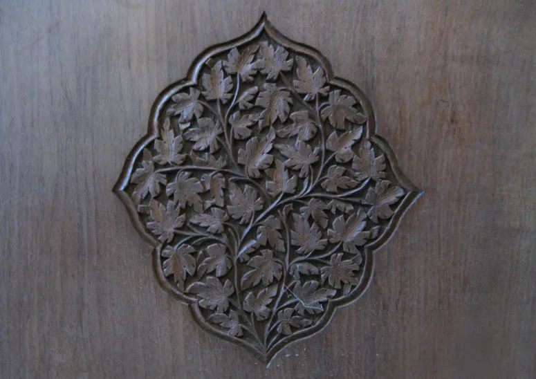 Close-up photograph of a Kashmiri walnut wood carver at work in a traditional workshop -- the carver is a middle-aged man with worn hands, seated cross-legged on a wooden platform, holding a short wooden-handled chisel in his right hand and a wooden mallet in his left, working on a large flat walnut panel (approximately 60cm wide) that rests on his lap and against a low wooden work bench, the panel shows a deep relief carving in progress -- a chinar leaf and arabesque vine design in naqqashi style, the background cut away 12-15mm deep with the design motifs standing proud in the warm brown walnut wood, fresh wood shavings and fine wood dust visible on the panel surface and on the carver's clothing, a shallow wooden tray holding an array of narrow gouges and flat chisels visible to the right, the workshop background is a rough stone wall with more finished carved panels leaning against it, natural light from a small window to the left giving warm directional illumination across the carving surface revealing the texture of the relief work