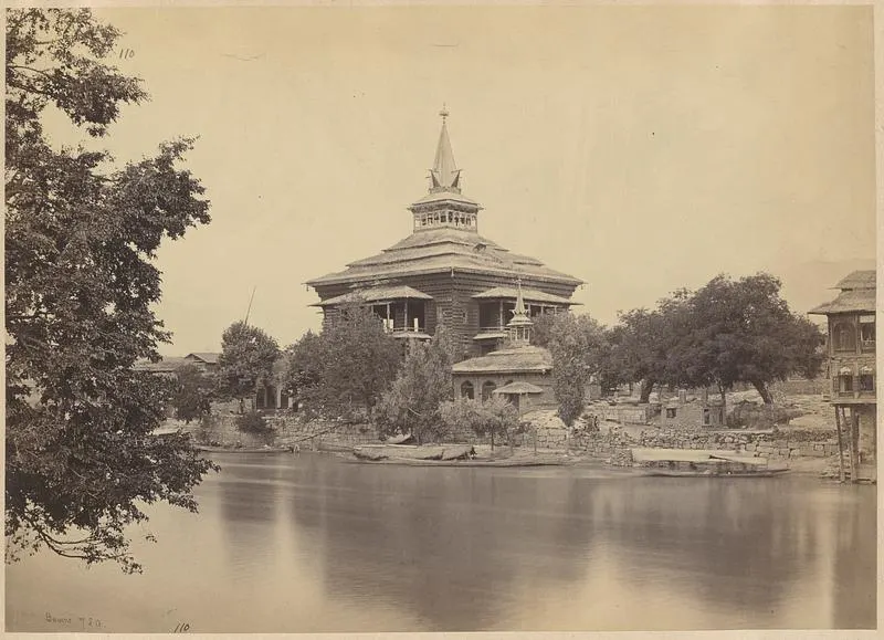 Wide-angle photograph of Khanqah Shah Hamdan from the Jhelum River bank in early October morning light -- the shrine's triple-tiered pyramidal roof in the traditional Kashmiri architectural style is fully visible above the river wall, the lowest tier of the roof widest with its swept wooden eaves, each successive tier narrower, finished with a copper finial at the apex, the exterior walls are covered in papier mache sheathing in intricate floral patterns painted in deep red, green and gold visible in the warm morning light, the Jhelum River in the foreground is grey-green and smooth at this morning hour with two wooden shikaras moored at the stone ghat steps below the shrine, the ghat steps are worn stone with a small wooden boat landing platform, the old city's four-storey timber-frame buildings visible on both sides of the shrine with their projecting wooden balconies, autumn light from the southeast at low angle giving long shadows and warm colour across the facade