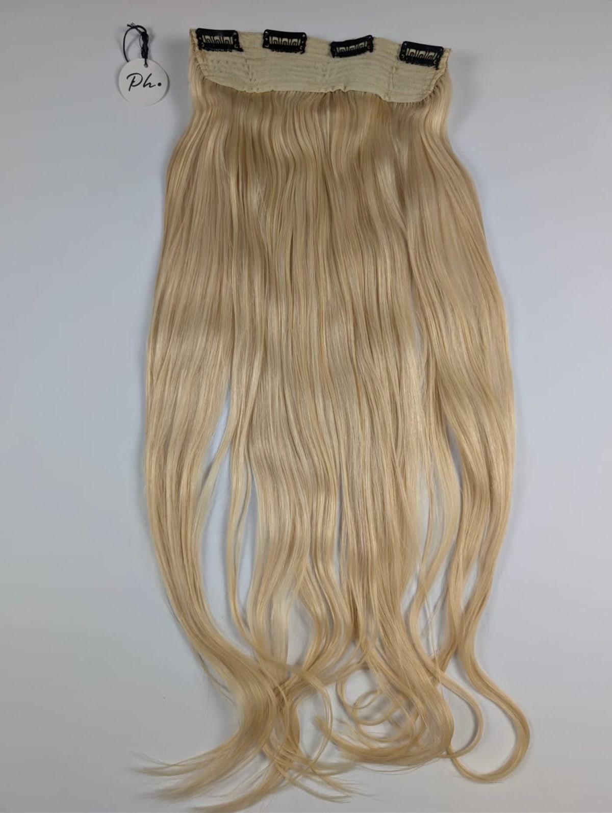 Long, wavy, blonde clip-in hair extension with four black clips, lying on a light gray surface, small tag attached.