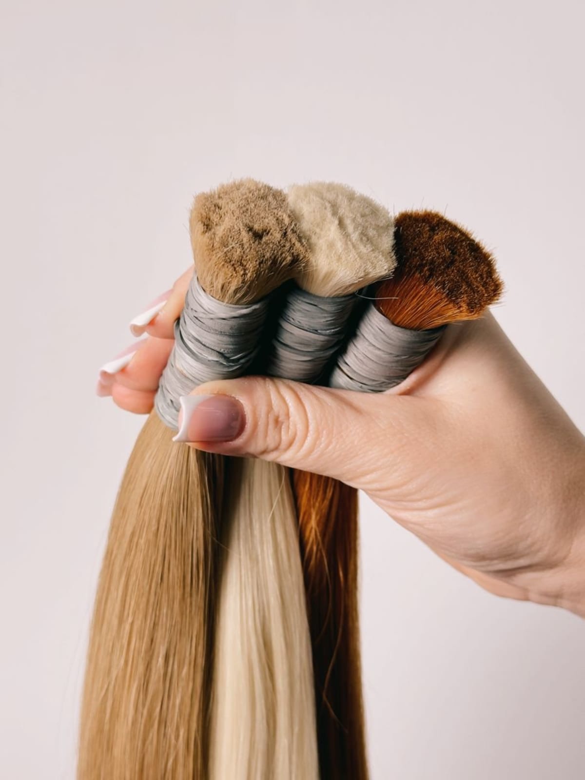 A hand holding three long straight hair extension wefts in blonde, light blonde, and auburn against a plain light background.