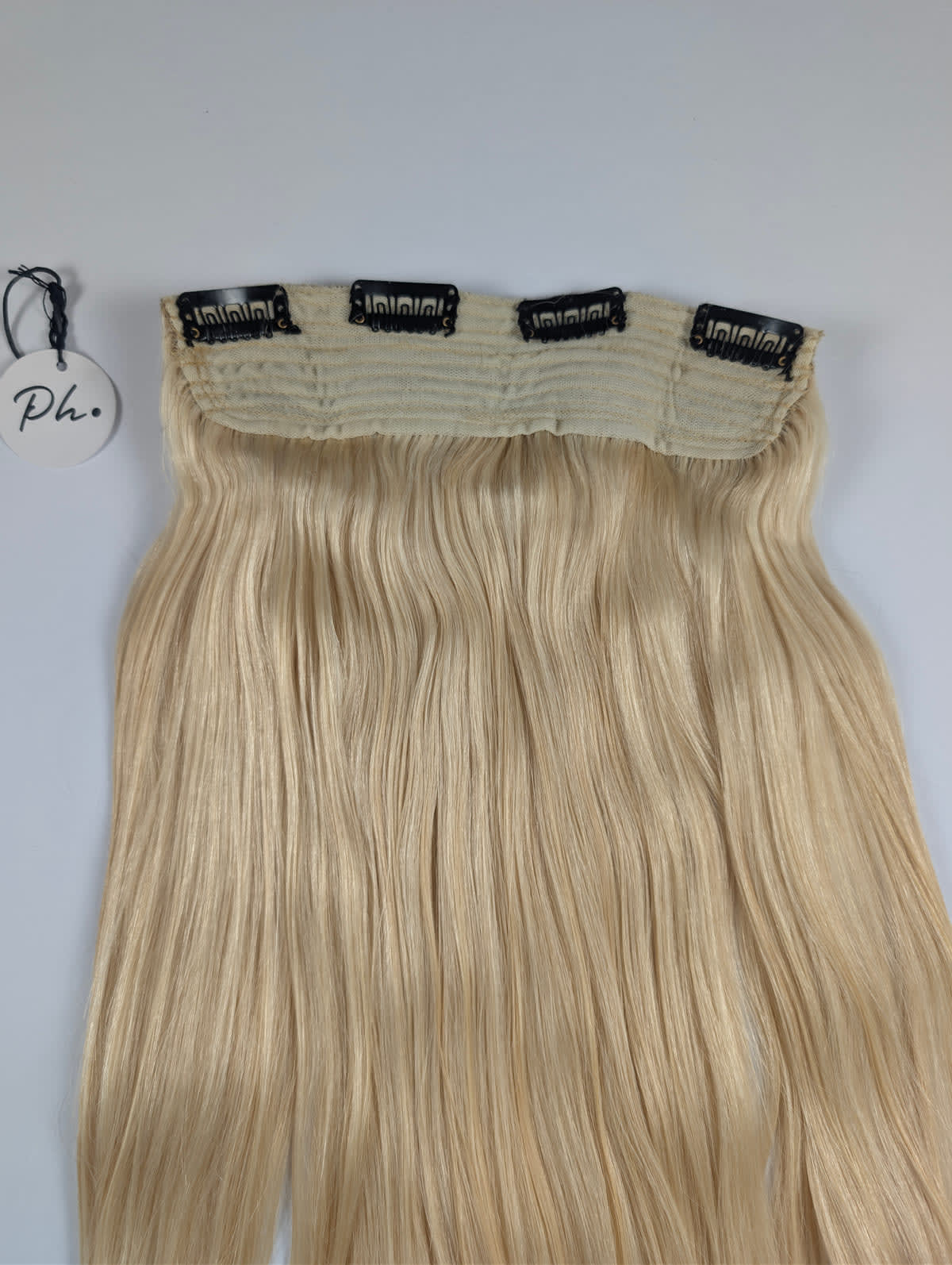 Extension di capelli biondi lunghi e lisci con quattro clip nere, su sfondo bianco, con un cartellino rotondo appeso.