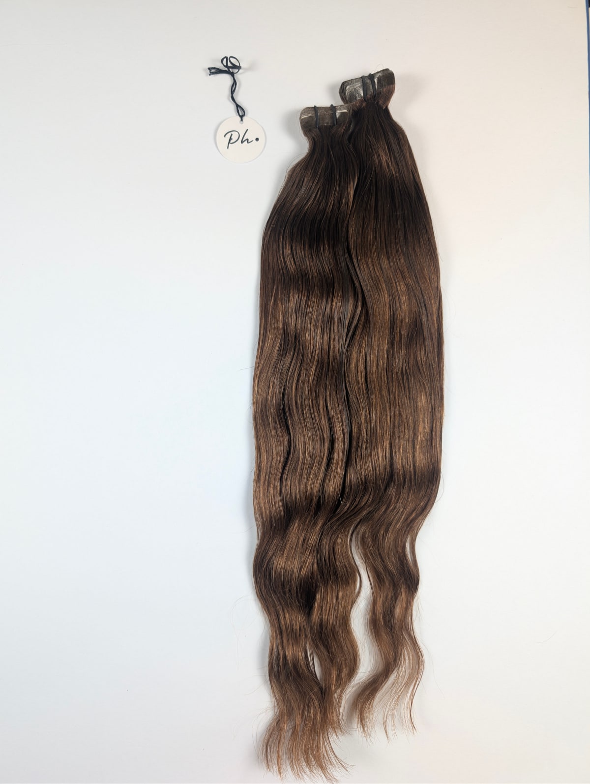 Zwei lange, braune, gewellte Haarsträhnen an Clips hängend an weißer Wand, daneben ein rundes Anhängerschild.