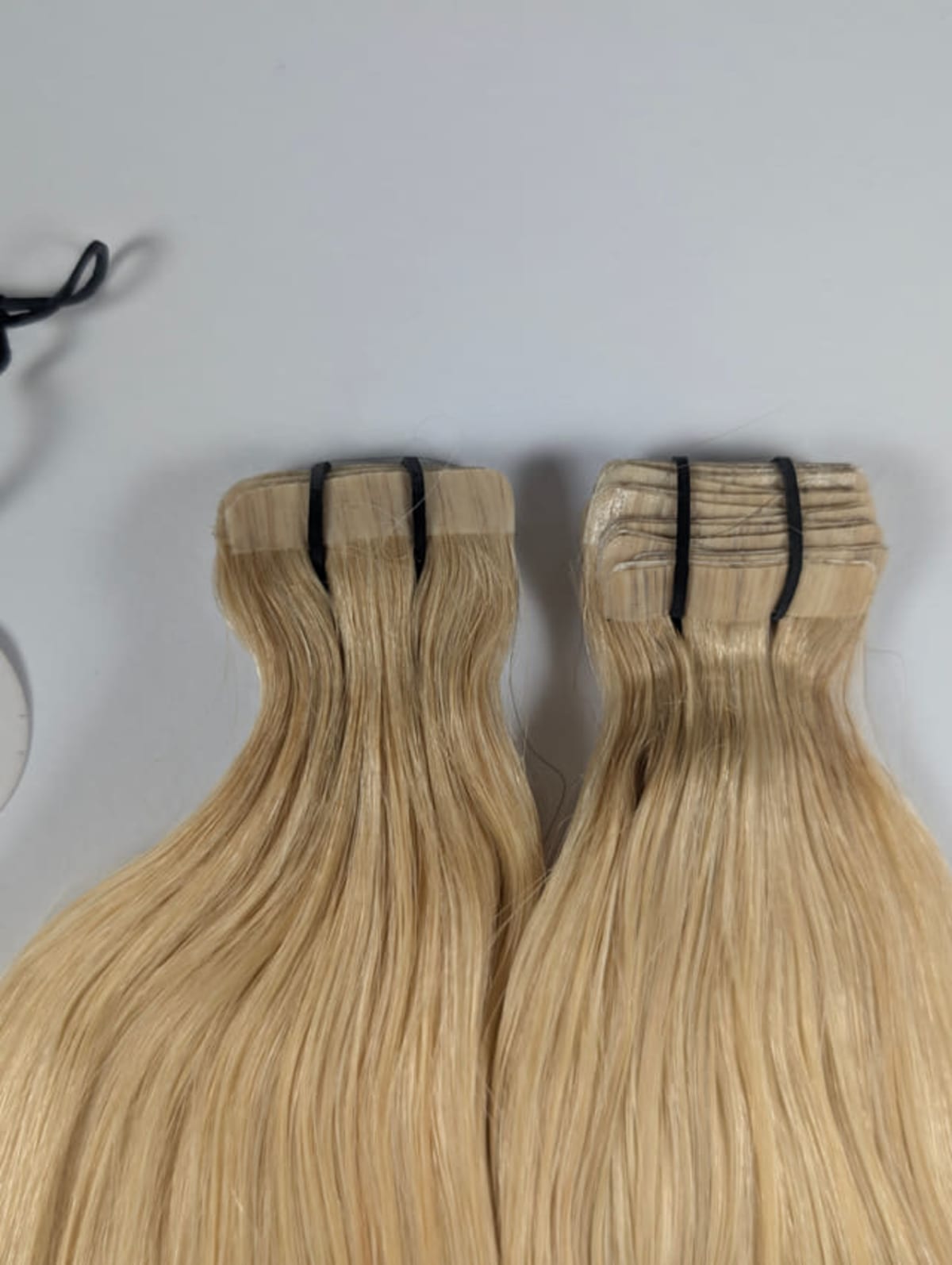 Zwei blonde, glatte Haarsträhnen mit schwarzen Bändern auf grauem Untergrund, links ein schwarzes Gummiband.