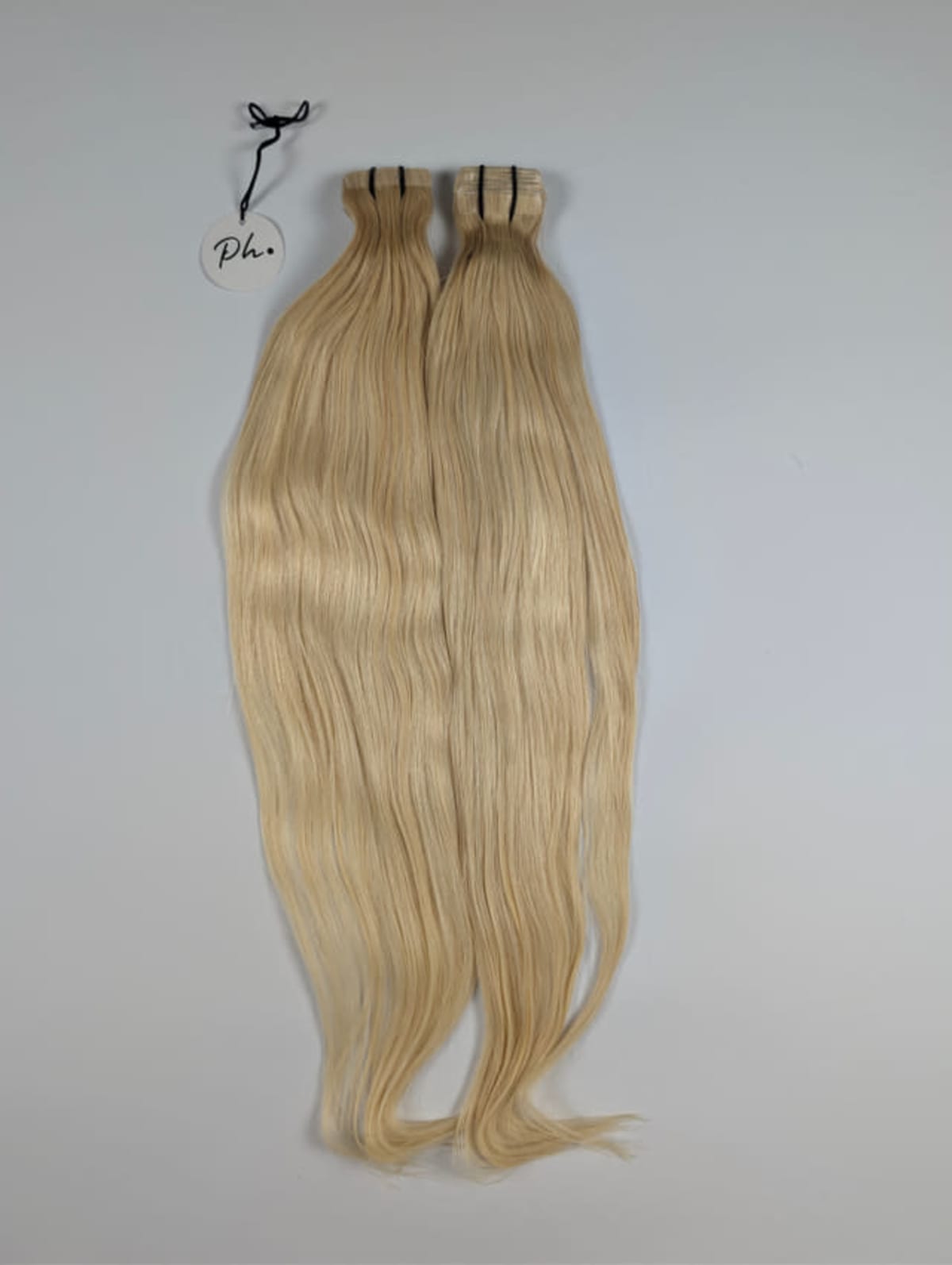Zwei lange, glatte, blonde Haartressen mit Etikett, liegend vor grauem Hintergrund.