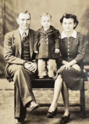 A black and white studio portrait from the 1940s: Tom Waters as a toddler standing on a bench between his mother and father.