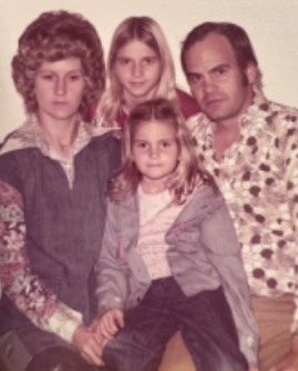 Tom and Brenda Waters seated together on a couch in the 1970s, Tom holding an acoustic guitar.
