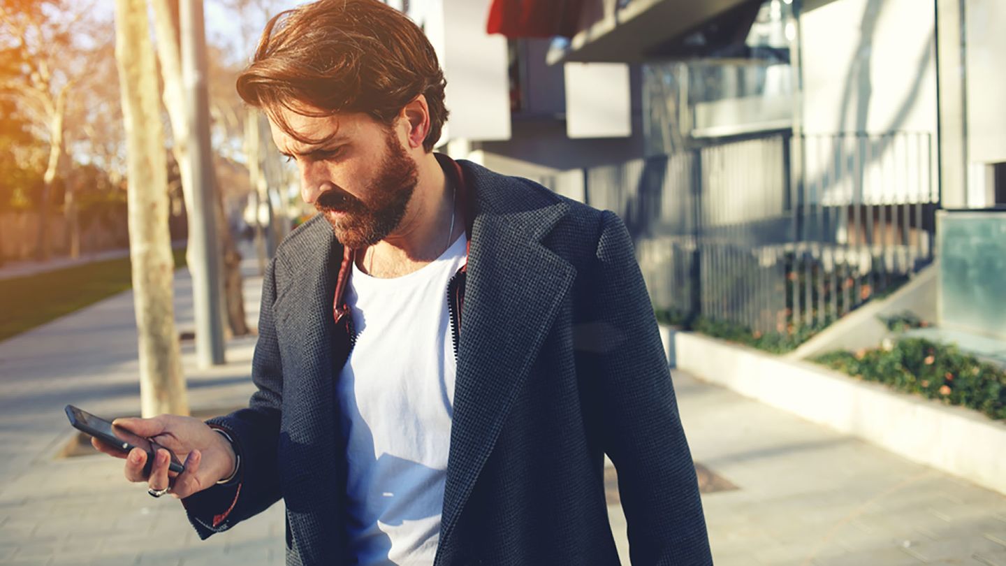 Bearded man walking on the street holding mobile whilst researching the internet for early signs of genital warts