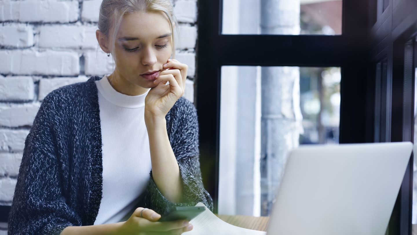 Woman sat in front of a window and a laptop checking on her phone if she has chronic cystitis