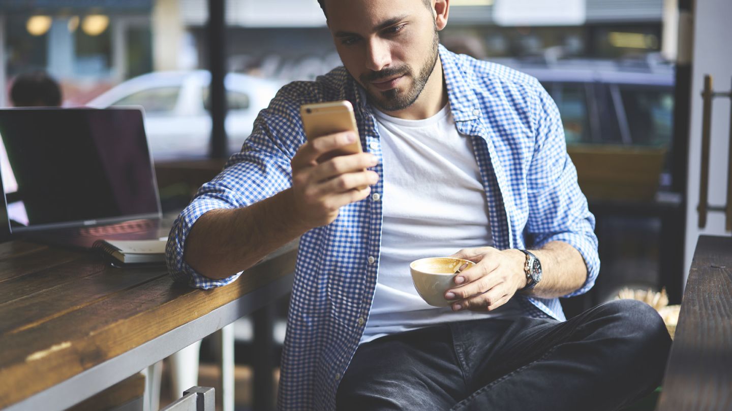 Man sat in a cafe holding a coffee cup checks for symptoms of chlamydia in men on his phone