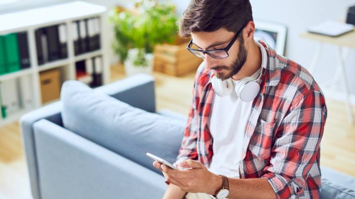 Caucasian man sat on back of couch at home staring at mobile phone researching signs of genital herpes