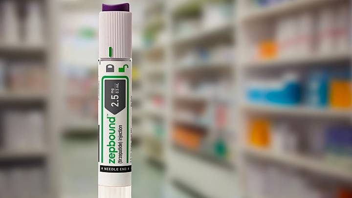 Un stylo injecteur Zepbound (tirz&eacute;patide) de 2,5 mg est pr&eacute;sent&eacute; debout devant un fond flou d&rsquo;&eacute;tag&egrave;res de pharmacie.