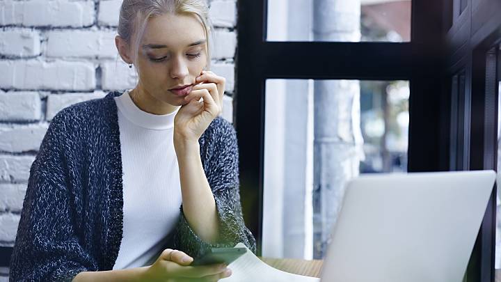Woman sat in front of a window and a laptop checking on her phone if she has chronic cystitis