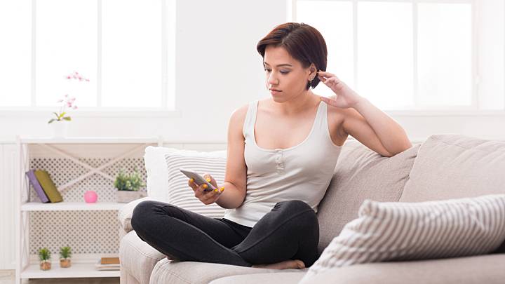Woman with short hair sat on couch at home looking at mobile phone researching causes of genital warts