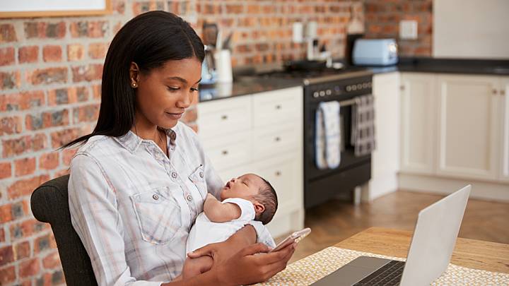 Woman sat in her kitchen in front of her laptop holding a baby is reading about contraception after giving birth on her phone