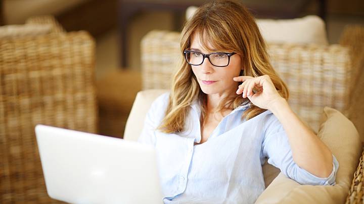 Woman with glasses sat in lounge chair looking up HRT side effects on her laptop