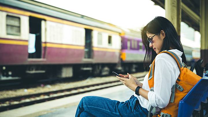 Eine Frau sitzt an einem Bahngleis. Sie informiert sich nach ihrer Reise über Malaria Symptome auf ihrem Handy.