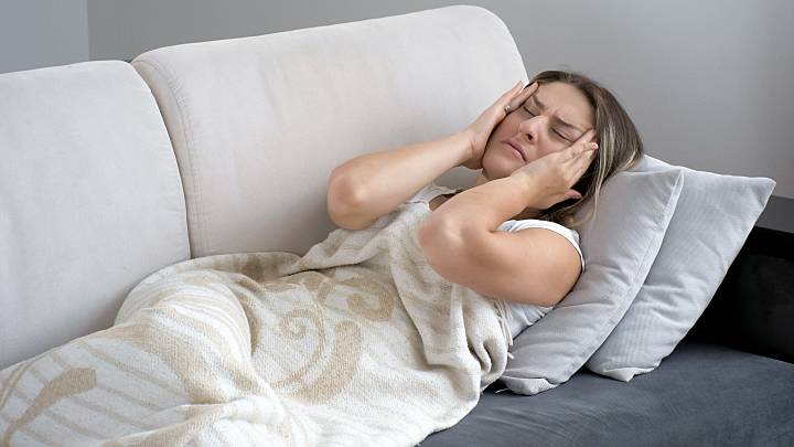 Eine Frau leidet an einem Migräneanfall. Sie liegt mit Kopfschmerzen auf ihrem Sofa und hält sich beide Hände auf ihren Kopf.