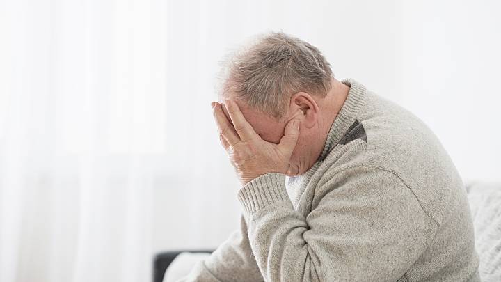 Bluthochdruck Symptome: Älterer Mann mit beigem Stickpullover sitzt auf der Couch und hält sich mit einer Hand den Kopf.