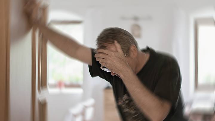 Hirninfarkt: Älterer Herr sieht verschwommen, hält sich mit einer Hand an den Kopf und stützt sich mit der anderen Hand an der Türe ab.