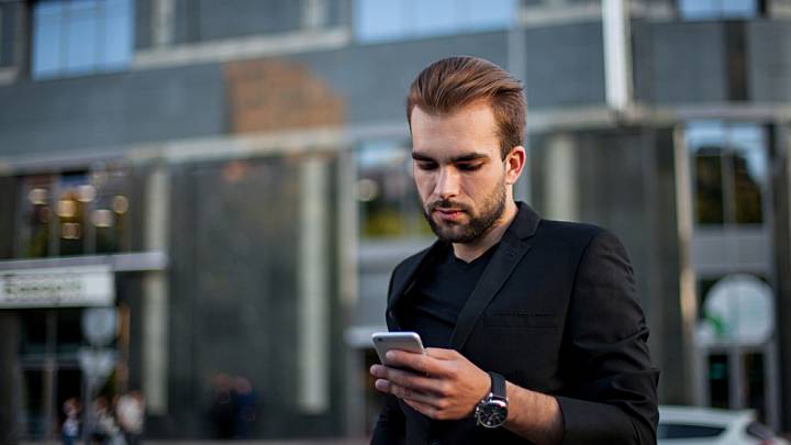 Man in black suit walking in city looking down at phone searching the internet for ways of preventing genital herpes