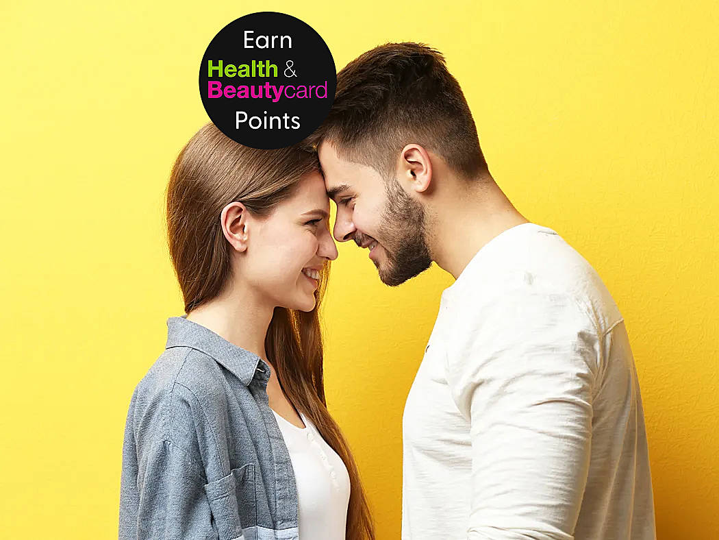 A young white man and woman who both have brown hair stand with their heads together smiling in front of a yellow background