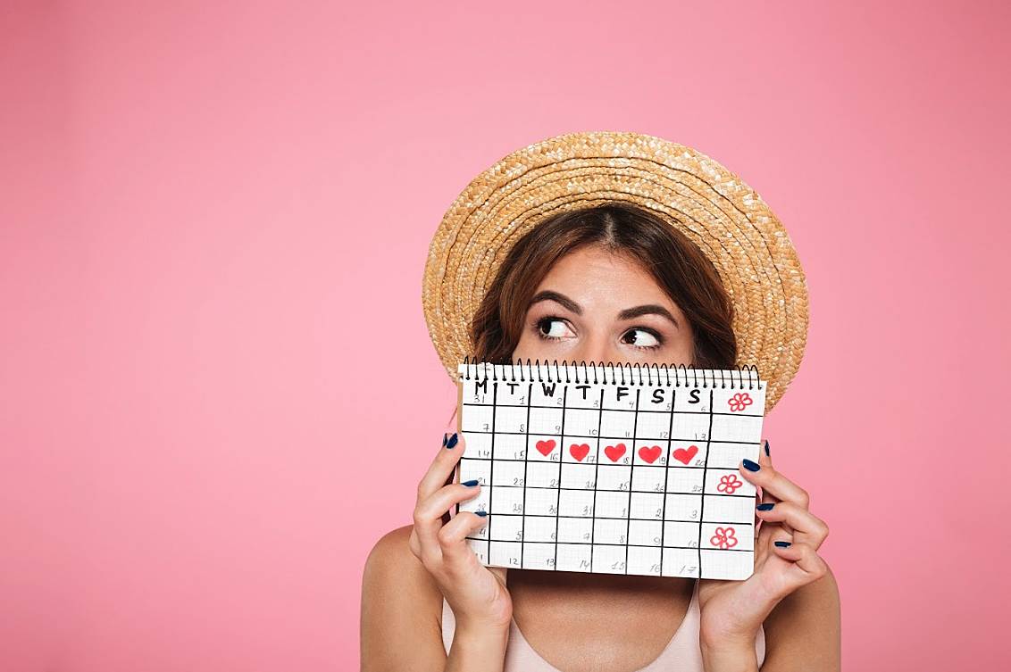 Woman with her holiday straw hat on holding up a calendar showing when she's planning to delay her period using treatment from Superdrug Online Doctor.