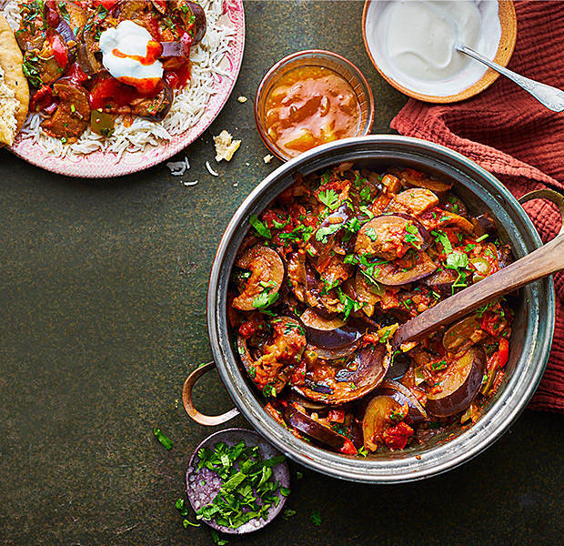 pan of aubergine curry being served into plate with condiments placed in bowls beside the plate