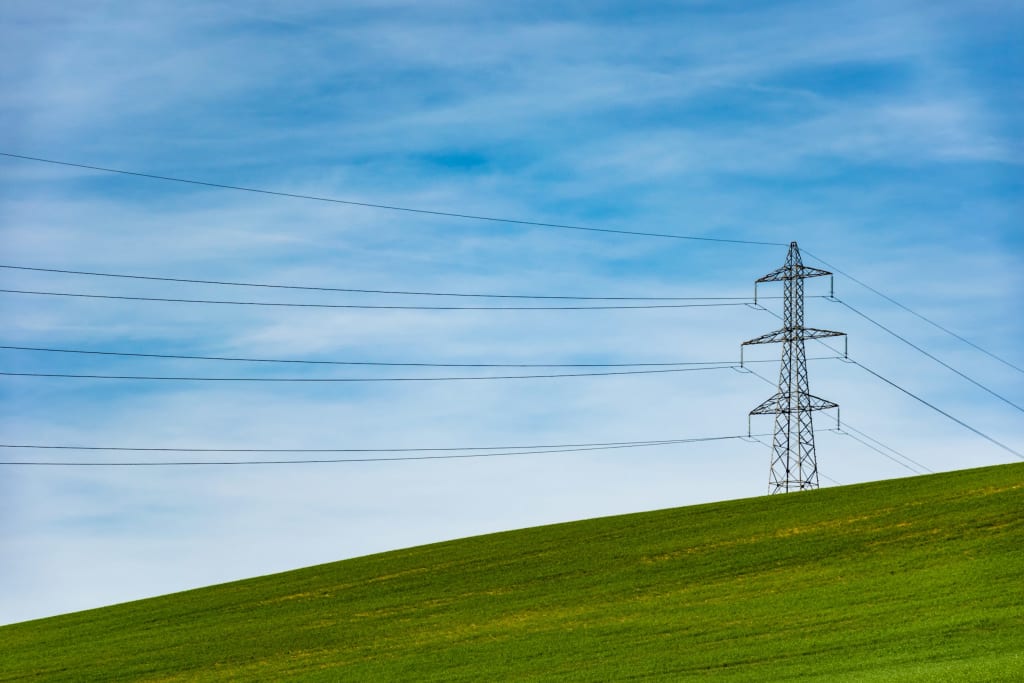 A lattice transmission tower on open ground carrying conductors across a hillside.