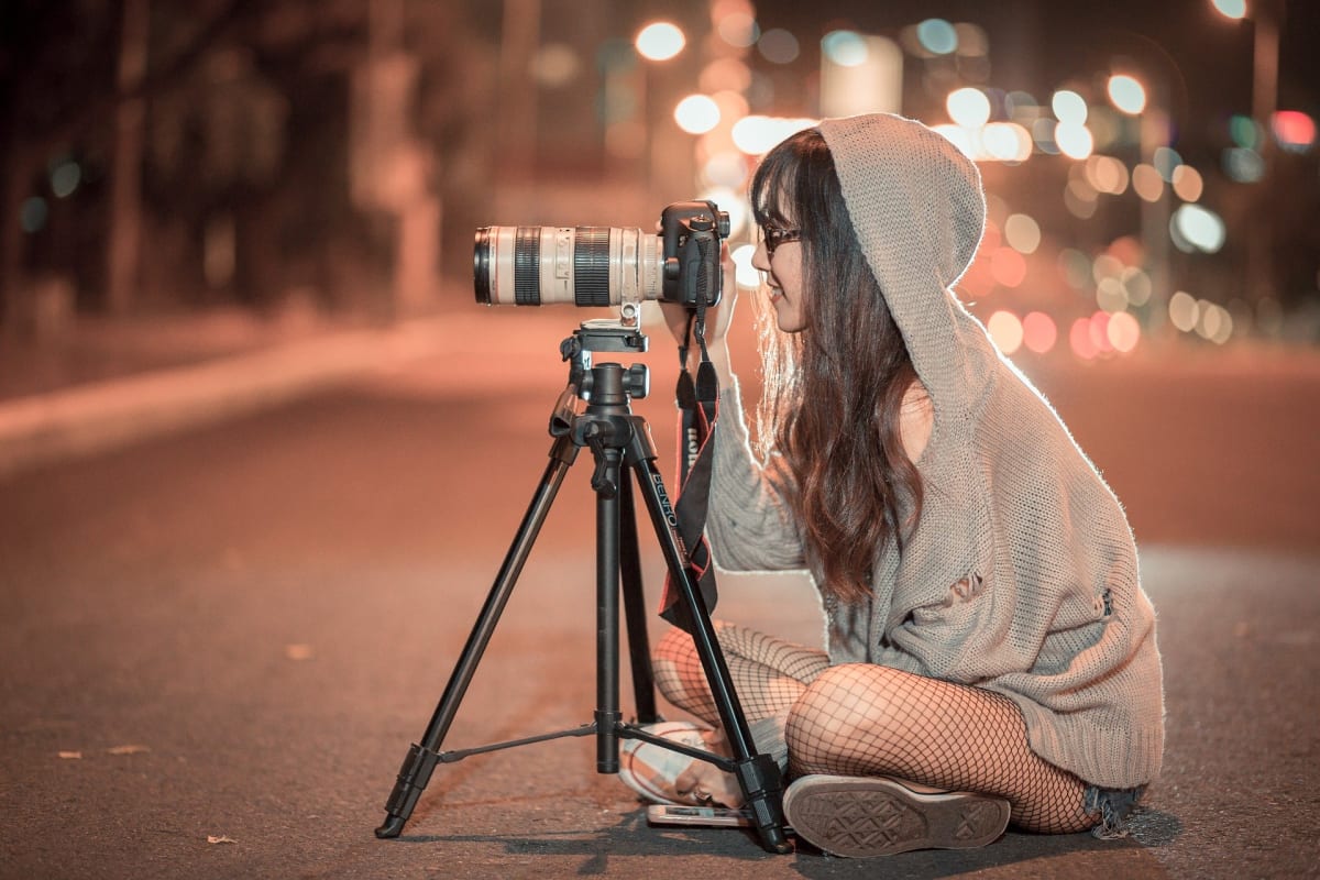 A woman sits cross-legged on the street behind a professional camera taking a photo.