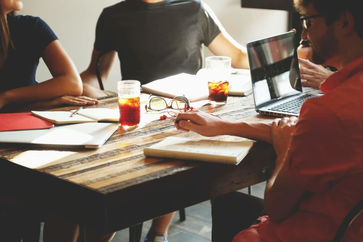 A group of people sit around a table covered with notebooks, a laptop, and some drinks.