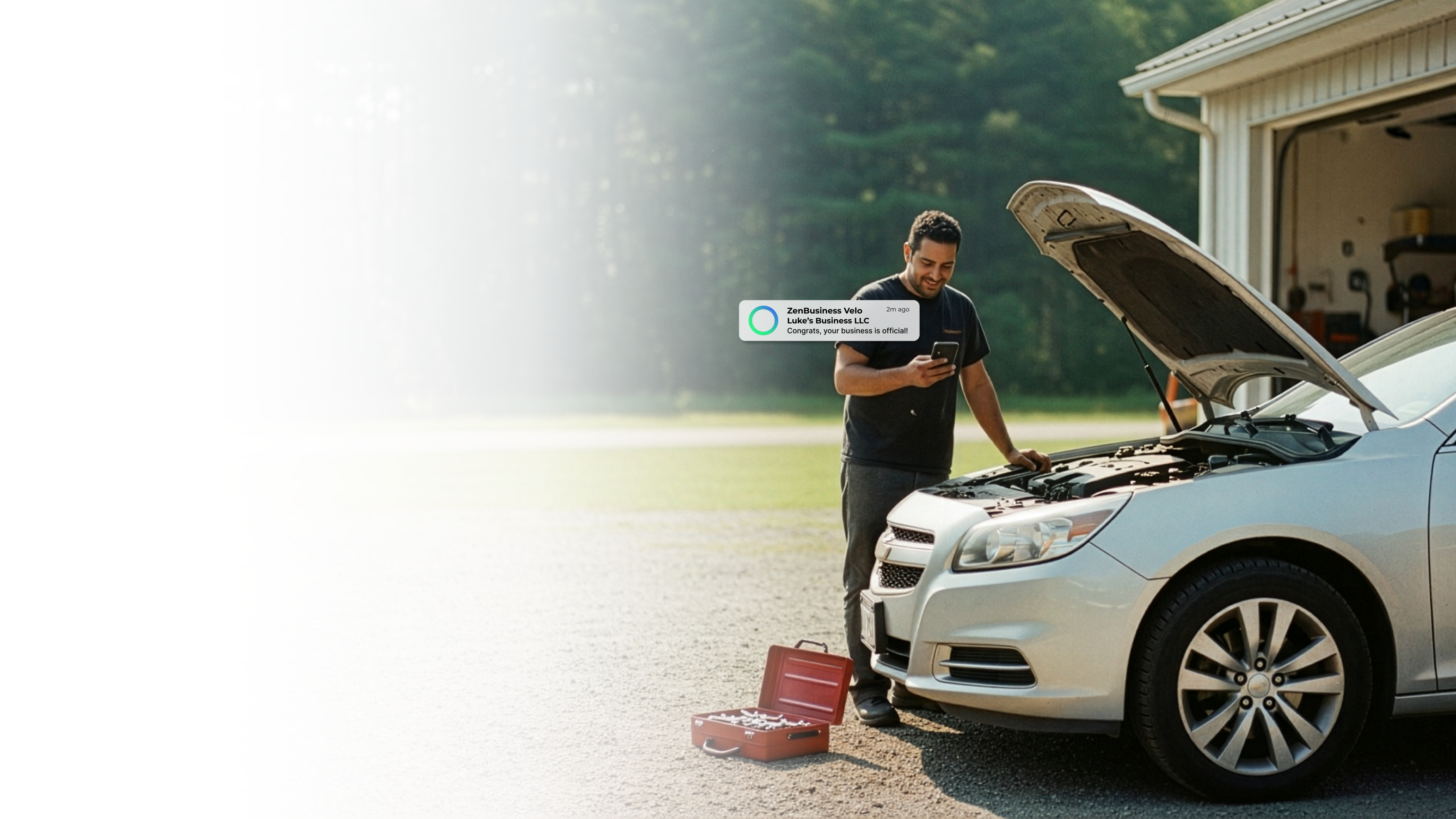 Male auto mechanic in Michigan smiling at his phone after ZenBusiness Velo confirmed his LLC is official while leaning over the hood of a car with the hood raised