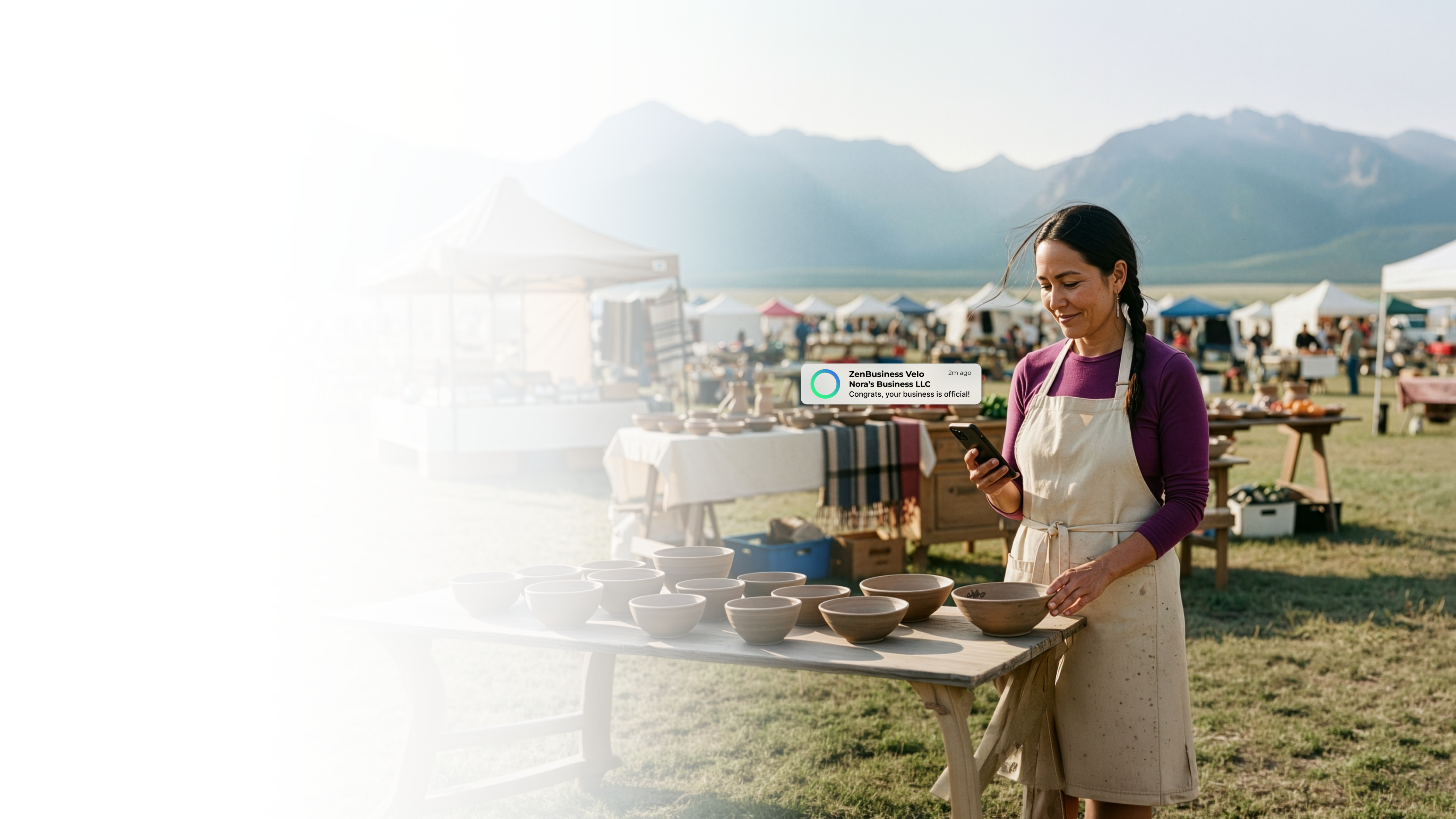 Female artisan vendor at an outdoor Montana market with handmade ceramic bowls on the table reading her ZenBusiness Velo notification confirming her LLC with mountain ranges in the background