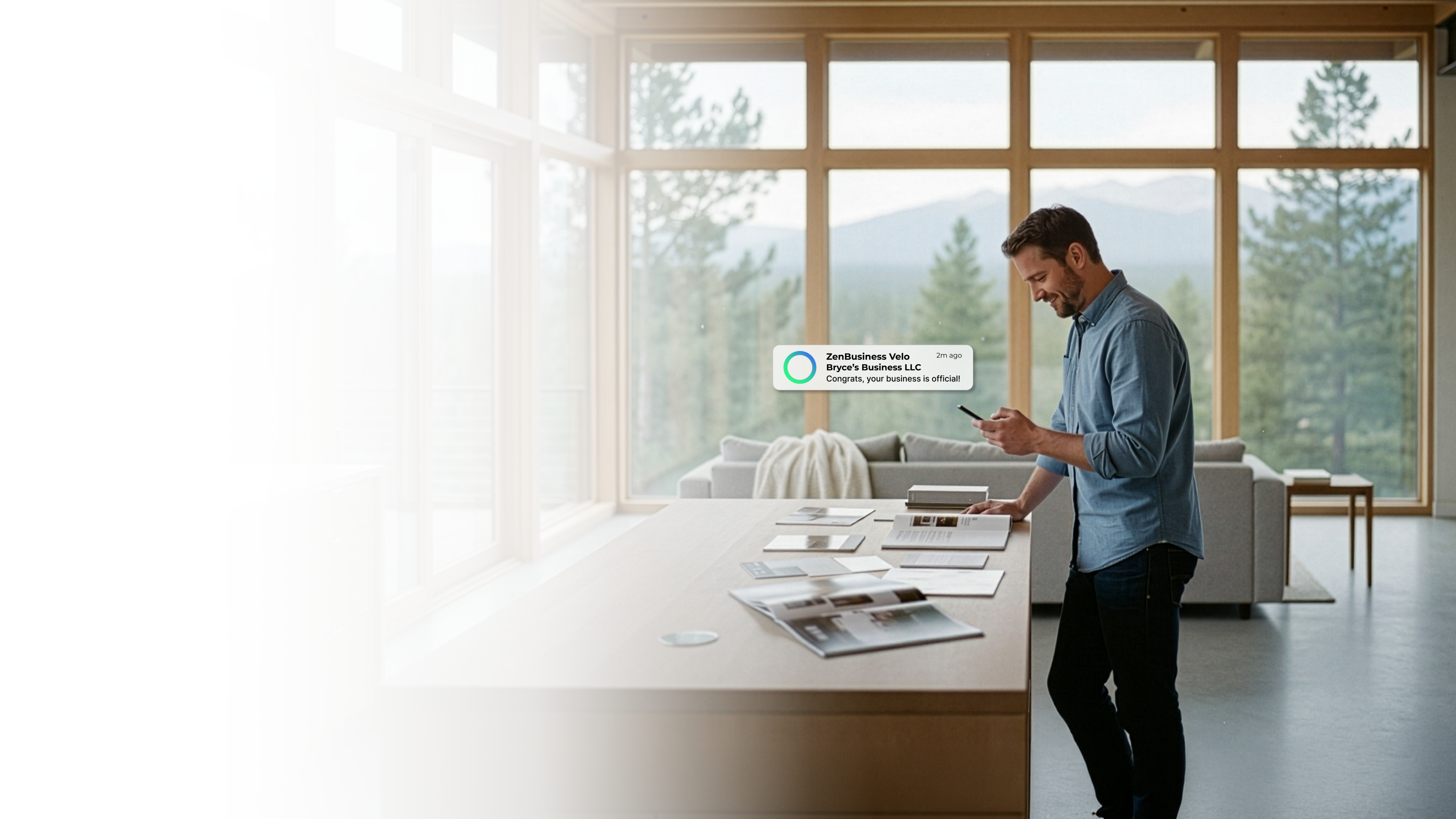 Male entrepreneur in a modern Colorado home studio with floor-to-ceiling windows and a pine forest view checking his LLC approval from a ZenBusiness Velo notification