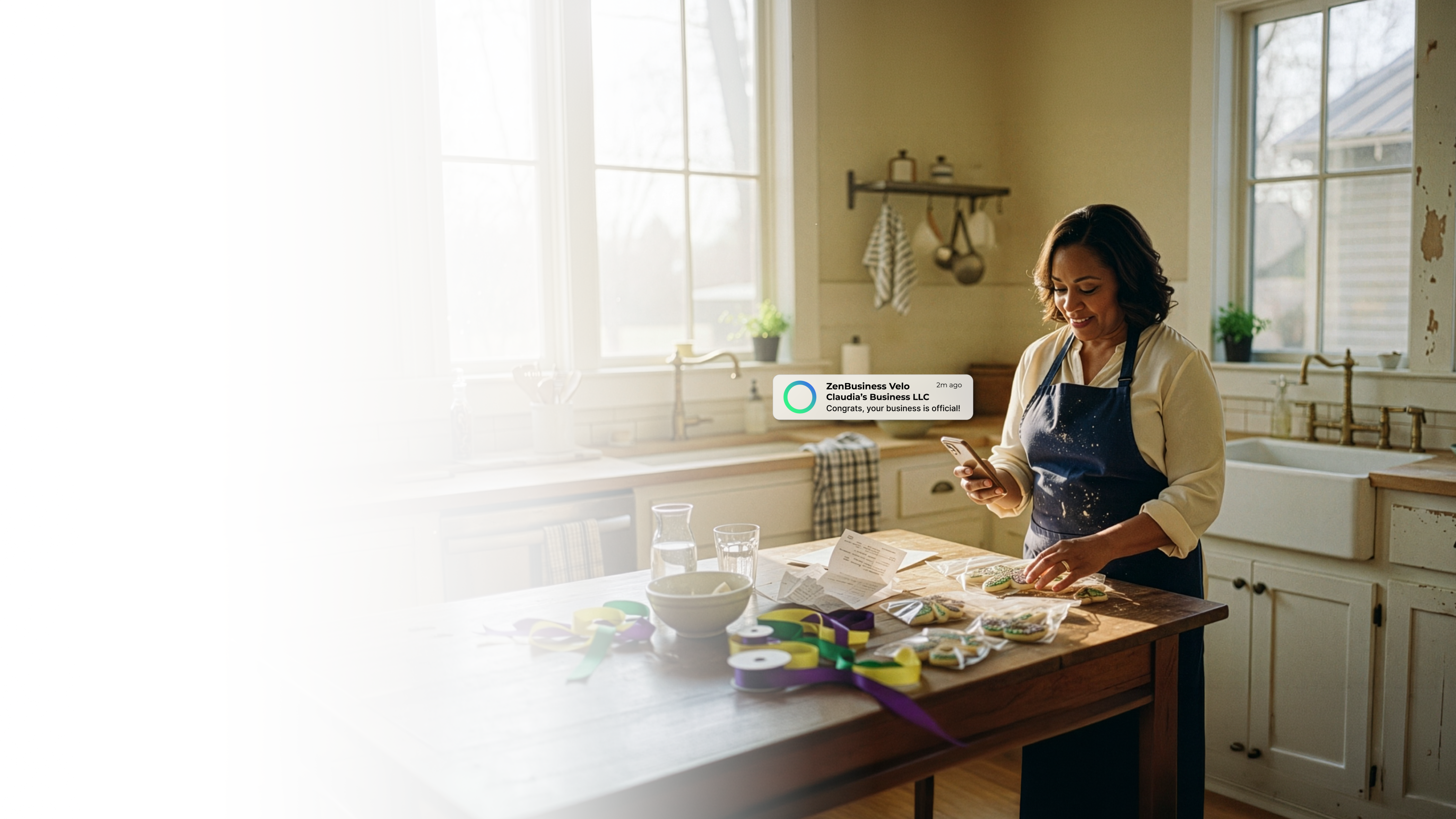 Female baker in a New Orleans Louisiana kitchen with Mardi Gras ribbons and decorated cookies on the counter checking her ZenBusiness Velo notification that her LLC is approved