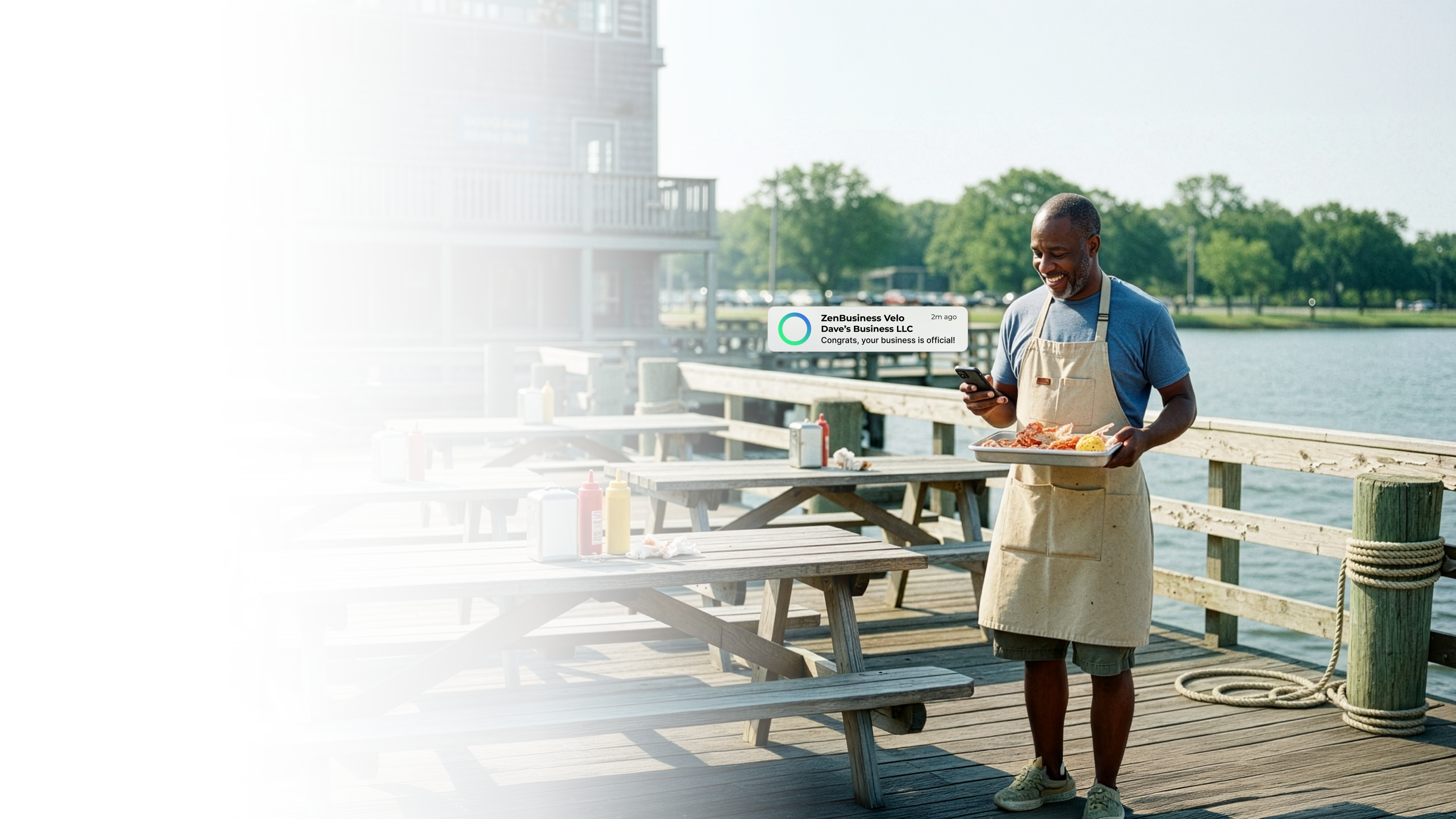 Male waterfront restaurant owner on a Maryland dock holding a food tray and checking his ZenBusiness Velo notification that his LLC is approved with a river and trees in the background