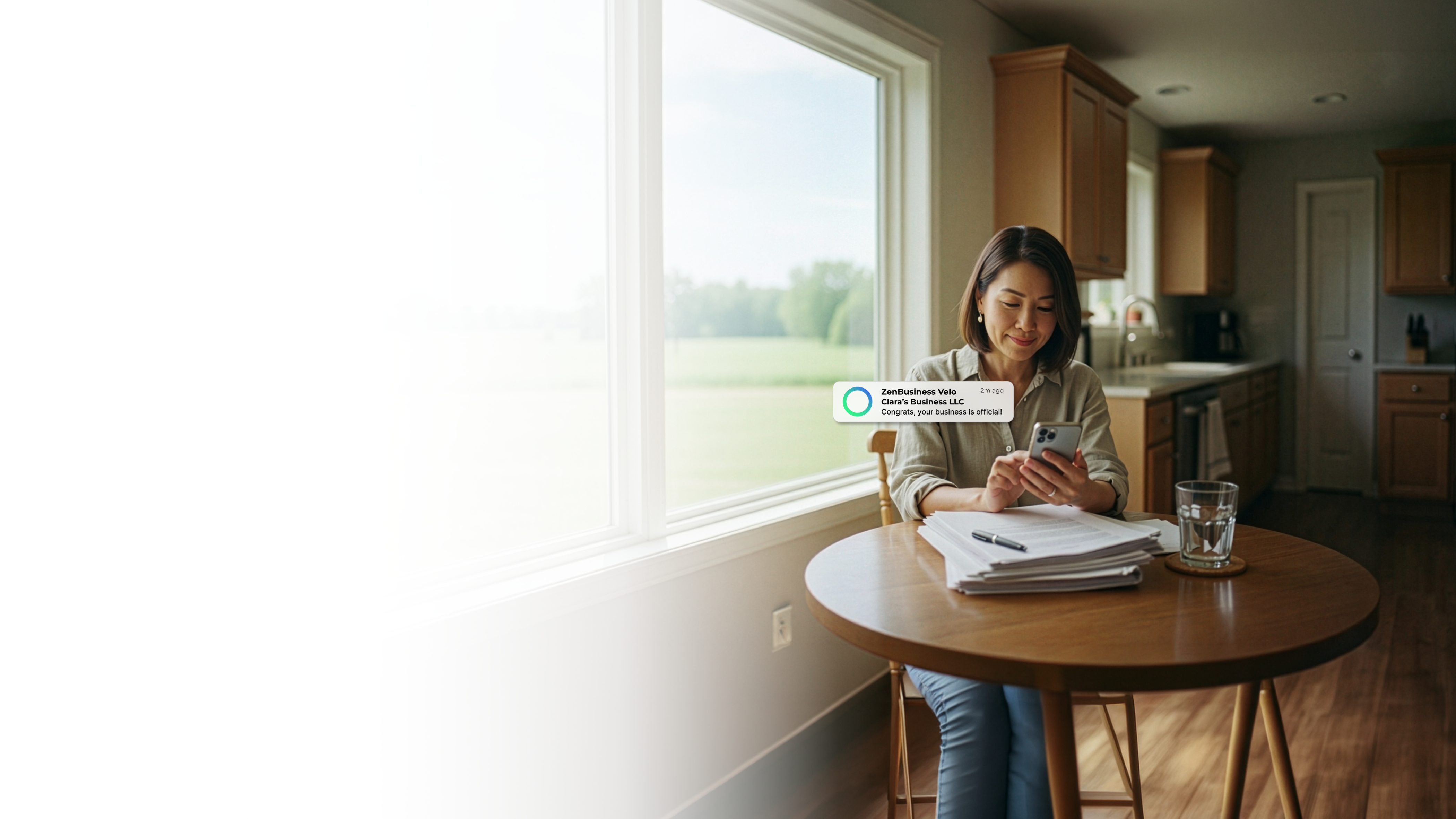 Female entrepreneur at a kitchen table in rural Minnesota with paperwork and a glass of water checking her ZenBusiness Velo notification confirming her LLC is official