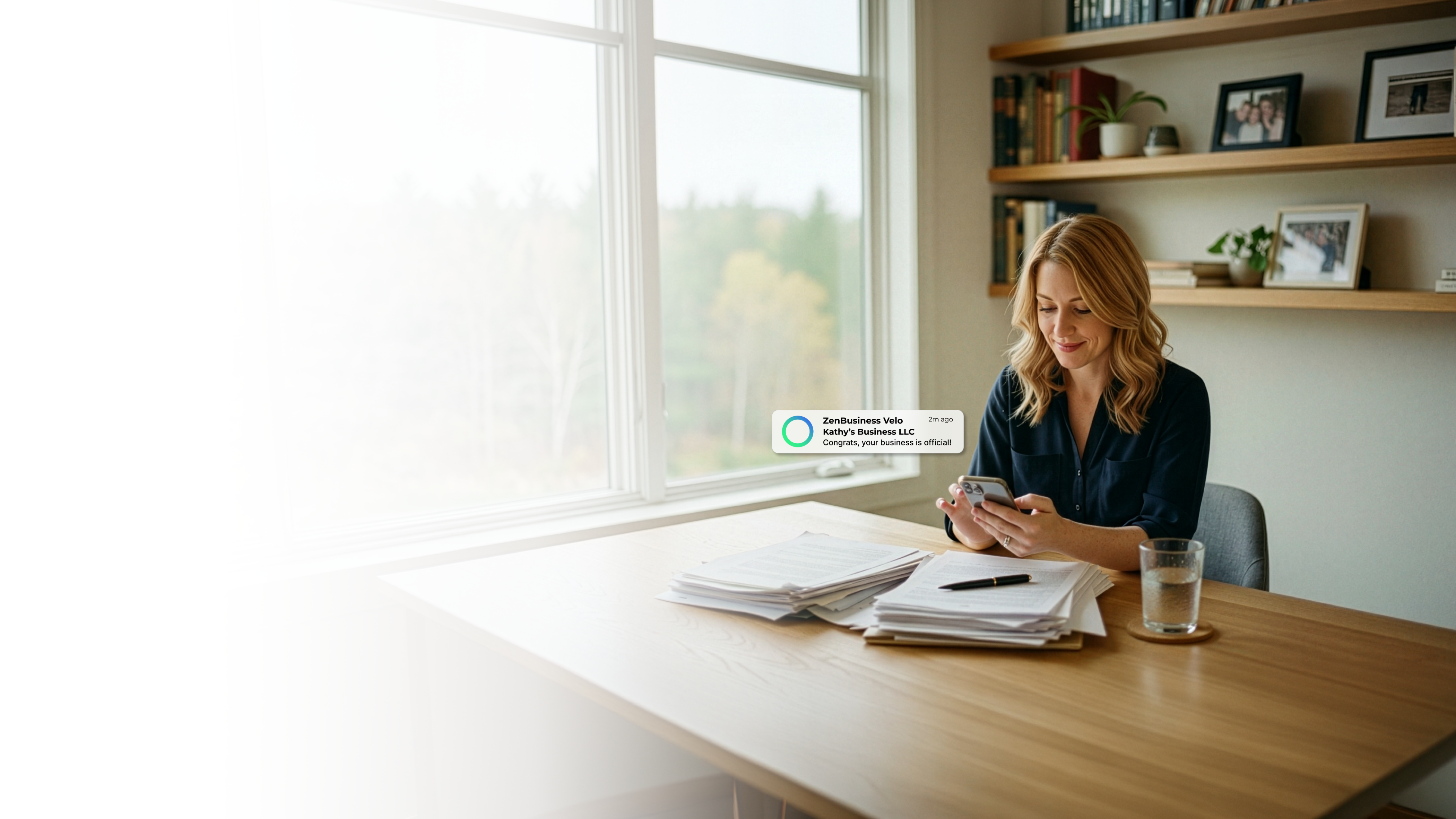 Female consultant at a home office desk in New Hampshire smiling at her ZenBusiness Velo notification that her LLC is official with bookshelves, paperwork, and a wooded autumn view through the window