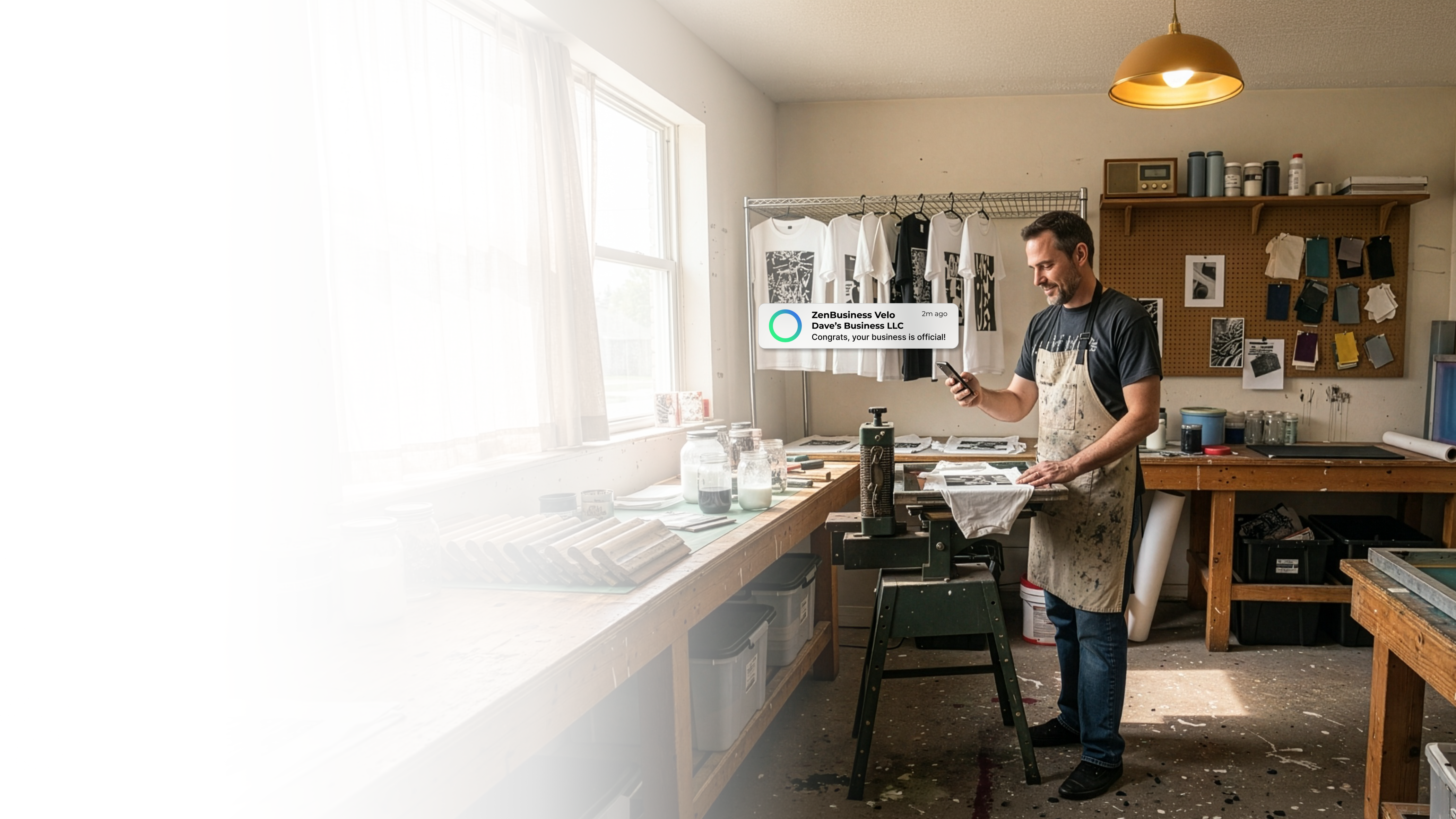 Male print shop owner in his Ohio studio with screen printing equipment and hanging t-shirts checking his phone after ZenBusiness Velo confirmed his LLC