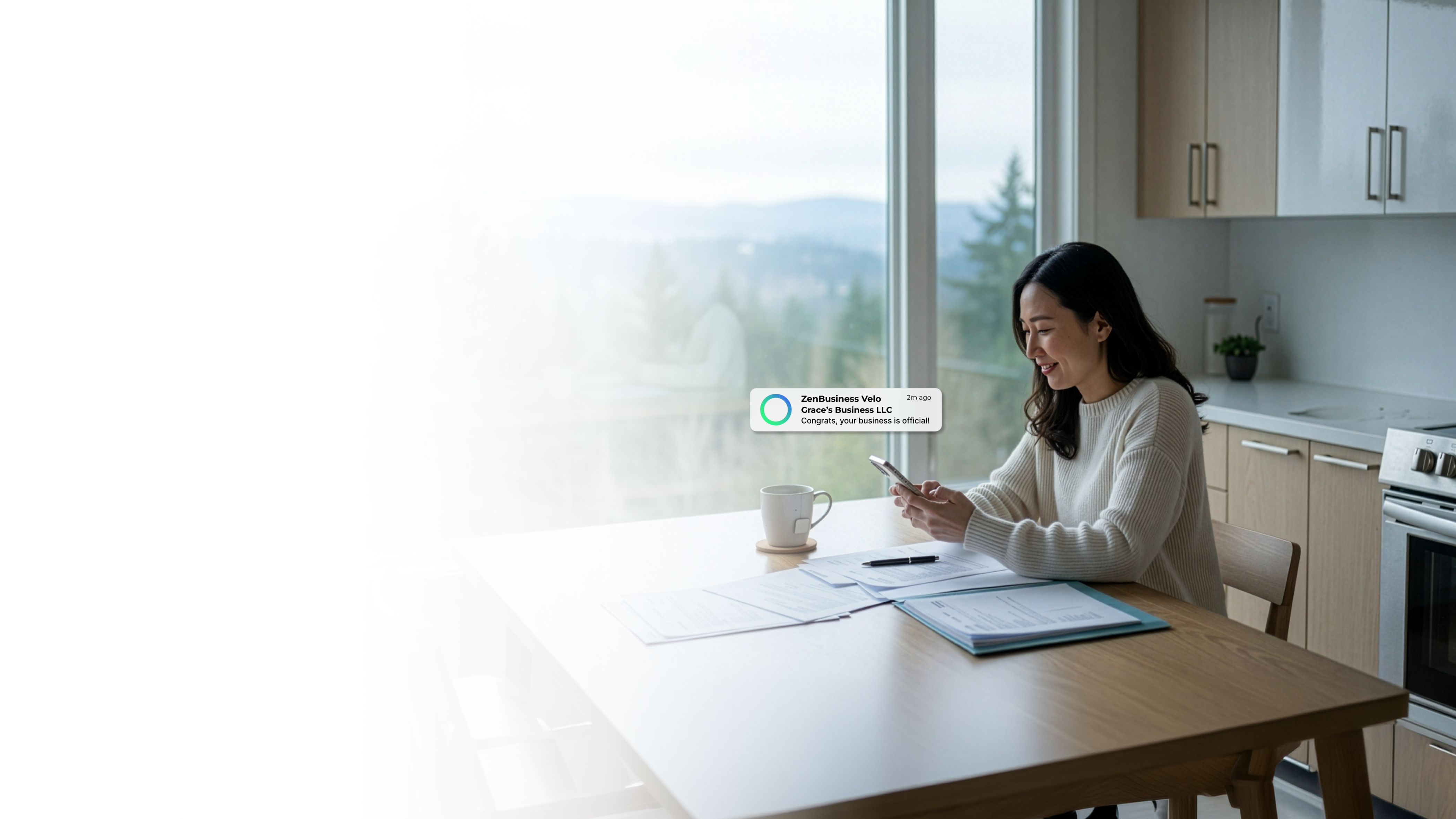 Female entrepreneur at a home office desk in Washington State reviewing paperwork and checking her ZenBusiness Velo notification that her LLC is official with a Pacific Northwest forest visible through the window